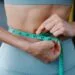 Person measuring waistline with a tape measure, representing weight loss progress and goals for the month.