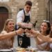 A group of five young adults (all over the legal drinking age of 18 in Italy), toast with wine glasses at an outdoor Italian cafe.