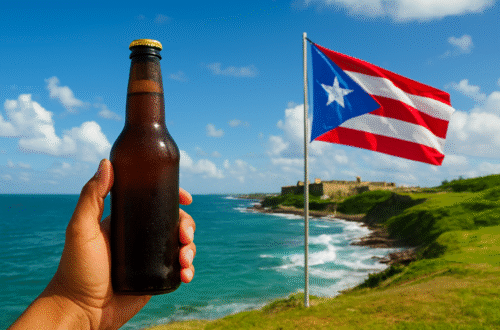 A person holding a cold beer bottle with the Puerto Rican flag waving near the ocean, representing Puerto Rico’s relaxed drinking age culture.