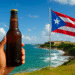 A person holding a cold beer bottle with the Puerto Rican flag waving near the ocean, representing Puerto Rico’s relaxed drinking age culture.