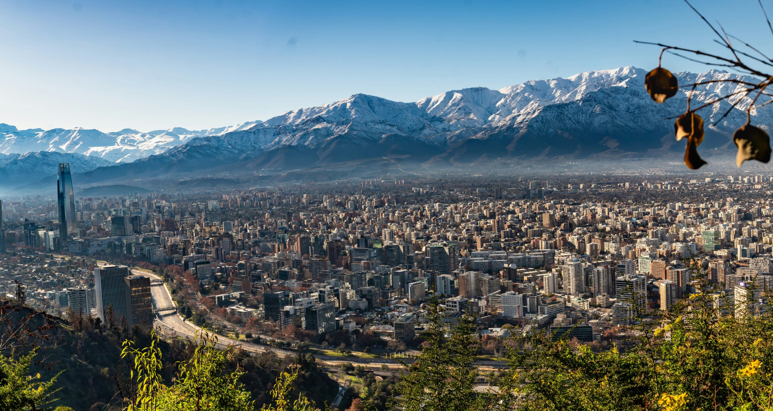 A panoramic view of Santiago, Chile, with the snow-capped Andes Mountains in the background. Lush greenery is visible in the foreground, and tall buildings are seen across the city's skyline.