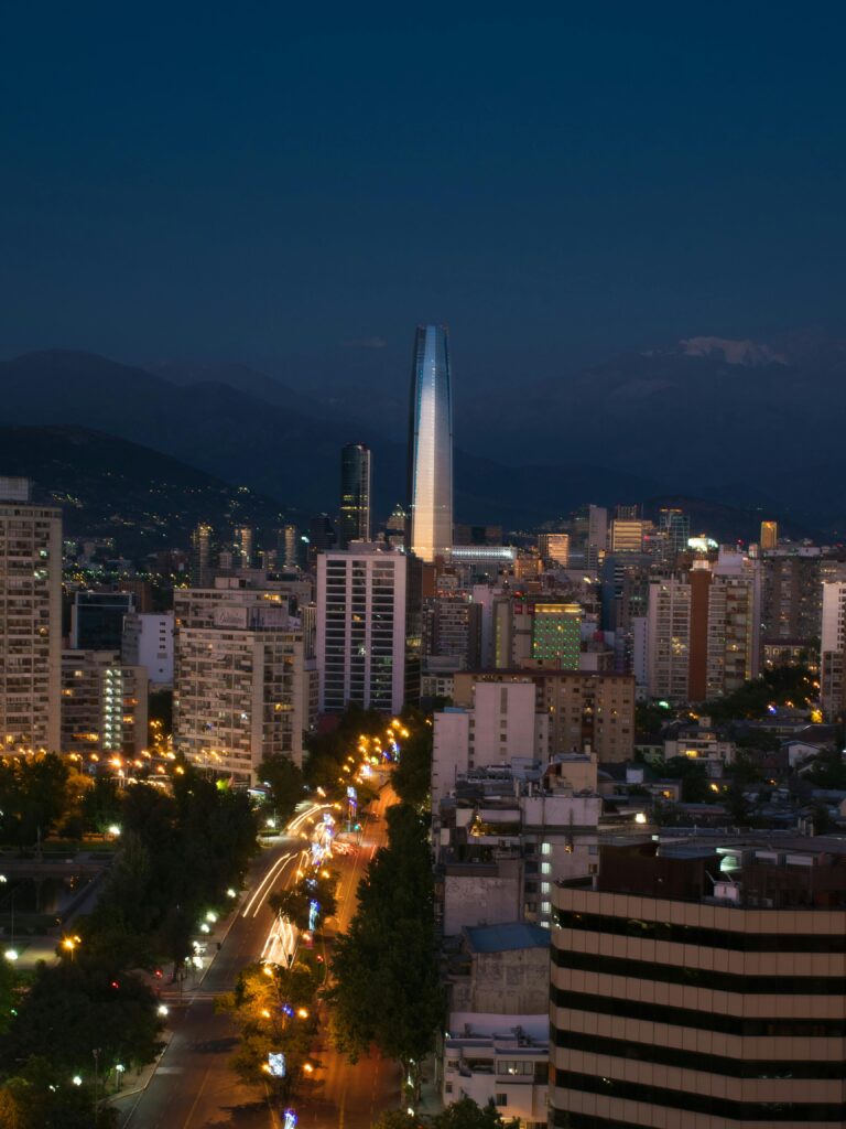 An elevated night shot of the city of Santiago, Chile. The city skyline is illuminated, dominated by the Gran Torre Santiago skyscraper standing prominently in the center. In the foreground, a long road shows streaks of car lights, suggesting movement. The faint silhouette of the Andes mountain range is visible in the background.
