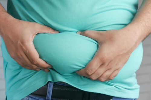 Close-up of a person wearing a light blue/green shirt and jeans, grabbing and pinching the excess skin and fat around their belly with both hands. Does Sweating Burn Belly Fat?