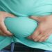 Close-up of a person wearing a light blue/green shirt and jeans, grabbing and pinching the excess skin and fat around their belly with both hands. Does Sweating Burn Belly Fat?