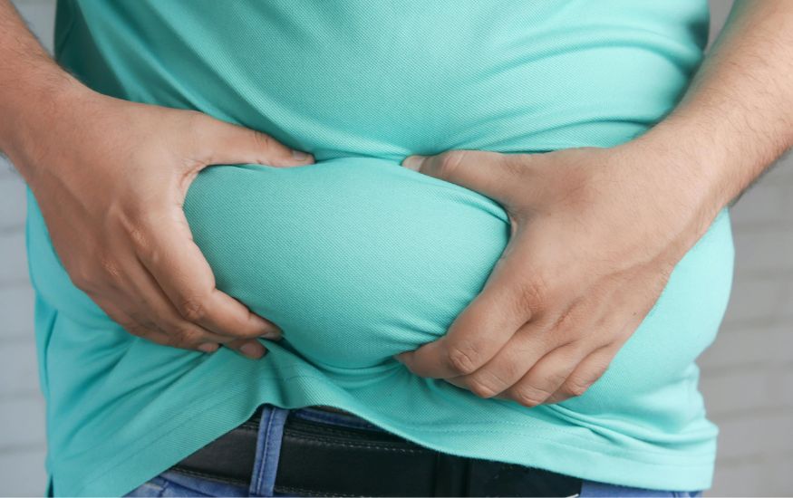 Close-up of a person wearing a light blue/green shirt and jeans, grabbing and pinching the excess skin and fat around their belly with both hands. Does Sweating Burn Belly Fat?