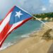 The Puerto Rican flag flying high over a beautiful, sunny tropical beach with turquoise water, golden sand, and a rugged cliffside in the background.