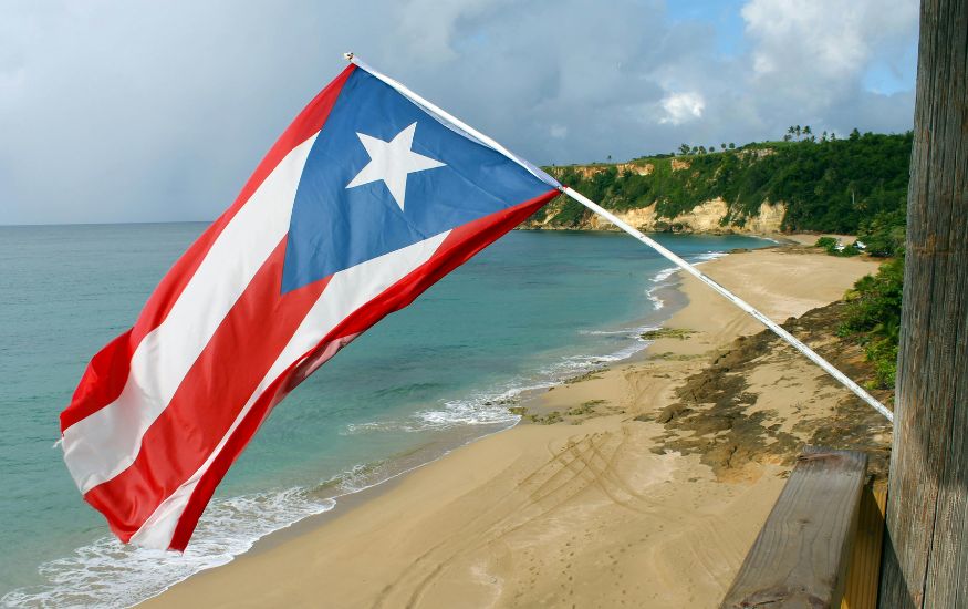 The Puerto Rican flag flying high over a beautiful, sunny tropical beach with turquoise water, golden sand, and a rugged cliffside in the background.