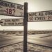 A vintage-style wooden signpost at a desert crossroads with three directional signs. One sign asks, "Is the Drinking Age 18 Anywhere in the United States?" while the others point toward US territories and the 50 states, indicating the legal age differences.