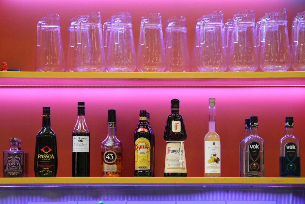 A magenta-lit bar shelf holds an array of liqueurs, including Kahlúa and a Puerto Rican spirit, beneath a row of clear glass pitchers.