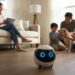 A small, spherical Ebo robot with glowing blue digital eyes sitting on a wooden floor in a sunlit living room, with a family and a dog interacting in the background.