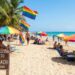 A sun-drenched photograph capturing a bustling gay beach scene. Rainbow pride flags and multicolored umbrellas dot the sand where diverse people are relaxing in lounge chairs. In the foreground, a wooden sign reads "Welcome to Sunset Beach - All are Welcome," with palm trees and turquoise ocean waves in the background.