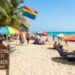 A sun-drenched photograph capturing a bustling gay beach scene. Rainbow pride flags and multicolored umbrellas dot the sand where diverse people are relaxing in lounge chairs. In the foreground, a wooden sign reads "Welcome to Sunset Beach - All are Welcome," with palm trees and turquoise ocean waves in the background.