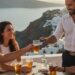 Glasses of Greek wine and Ouzo on a rustic wooden table at a seaside taverna, with white-washed buildings and the blue Mediterranean Sea in the background.drinking age in greece