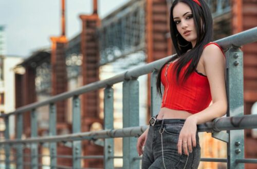 A woman with long black hair posing on an industrial bridge wearing a vibrant red zip-up top and charcoal skinny jeans with a silver chain belt. Are Skinny Jeans in Style 2026? This image showcases how the slim-fit silhouette remains a staple in edgy, urban streetwear.