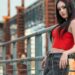 A woman with long black hair posing on an industrial bridge wearing a vibrant red zip-up top and charcoal skinny jeans with a silver chain belt. Are Skinny Jeans in Style 2026? This image showcases how the slim-fit silhouette remains a staple in edgy, urban streetwear.
