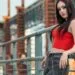 A woman with long black hair posing on an industrial bridge wearing a vibrant red zip-up top and charcoal skinny jeans with a silver chain belt. Are Skinny Jeans in Style 2026? This image showcases how the slim-fit silhouette remains a staple in edgy, urban streetwear.