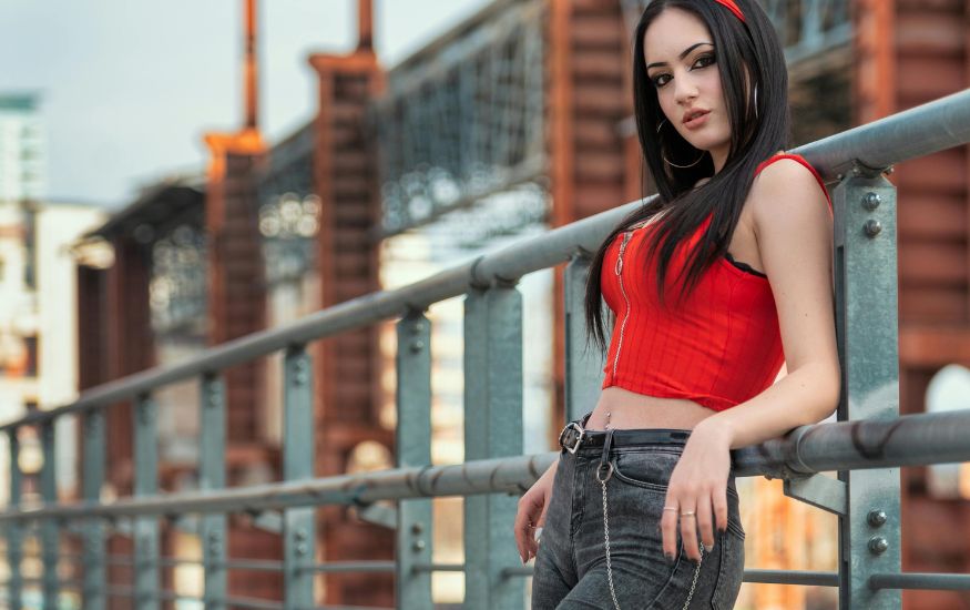 A woman with long black hair posing on an industrial bridge wearing a vibrant red zip-up top and charcoal skinny jeans with a silver chain belt. Are Skinny Jeans in Style 2026? This image showcases how the slim-fit silhouette remains a staple in edgy, urban streetwear.