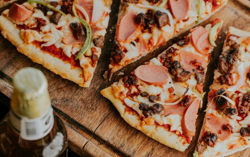 Slices of delicious Brooklyn Style Pizza topped with pepperoni, ham, sausage, and peppers on a rustic wooden board.