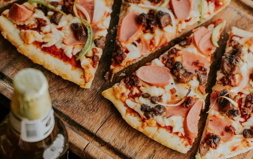 Slices of delicious Brooklyn Style Pizza topped with pepperoni, ham, sausage, and peppers on a rustic wooden board.