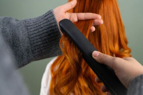 A close-up of a hair stylist using a flat iron on a section of vibrant copper-colored hair. What is silk press? It is a styling method used to get natural, curly, or textured hair into a sleekly styled, smooth, and shiny look without the use of chemical treatments.