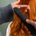 A close-up of a hair stylist using a flat iron on a section of vibrant copper-colored hair. What is silk press? It is a styling method used to get natural, curly, or textured hair into a sleekly styled, smooth, and shiny look without the use of chemical treatments.