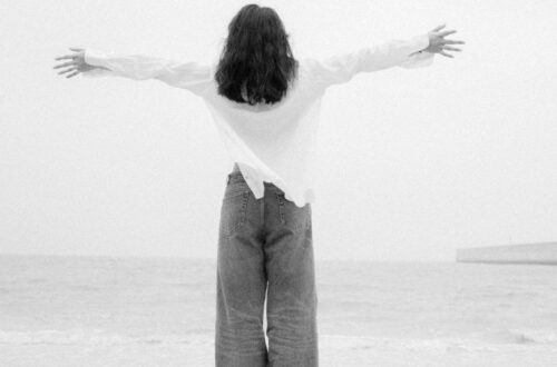 A black-and-white photograph of a person from behind with arms outstretched toward the ocean, wearing a relaxed white button-down shirt and denim trousers, demonstrating how to style wide leg jeans for a breezy, coastal look.