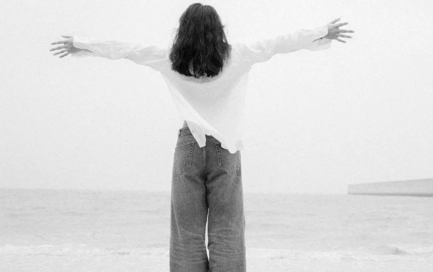 A black-and-white photograph of a person from behind with arms outstretched toward the ocean, wearing a relaxed white button-down shirt and denim trousers, demonstrating how to style wide leg jeans for a breezy, coastal look.