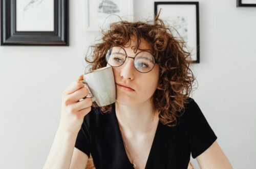 A young woman with round glasses and voluminous curls holding a coffee mug to her cheek, illustrating how to style short curly hair in a natural, messy-chic look.