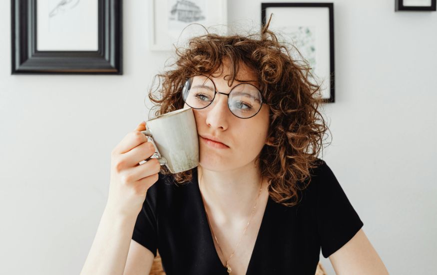 A young woman with round glasses and voluminous curls holding a coffee mug to her cheek, illustrating how to style short curly hair in a natural, messy-chic look.