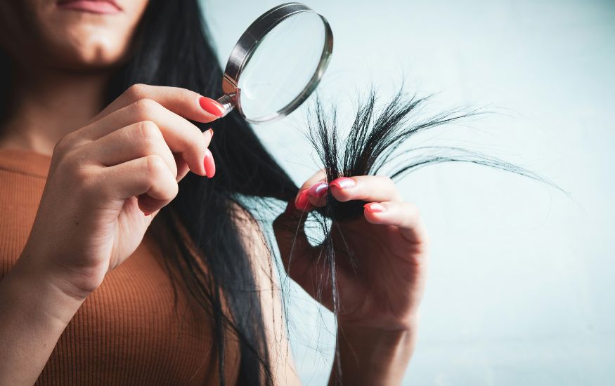 A close-up of a person using a magnifying glass to examine frayed split ends, showing exactly what Damaged Hair looks like under inspection.