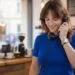 A young woman with a brown wolf cut hairstyle smiling while talking on a vintage black rotary phone in a cozy cafe setting. How to style a wolf cut to look effortless and voluminous.
