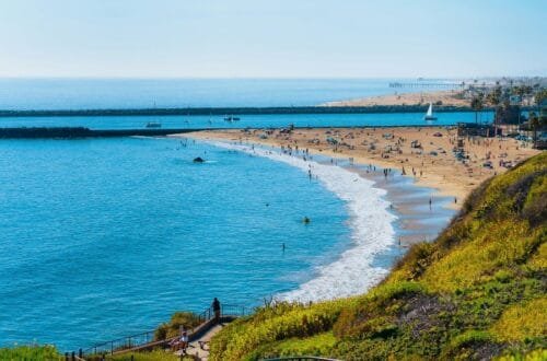 A scenic aerial view of a sunny coast showcasing popular things to do in Newport Beach, with beachgoers enjoying the clear ocean waters, sandy shore, and grassy overlook.