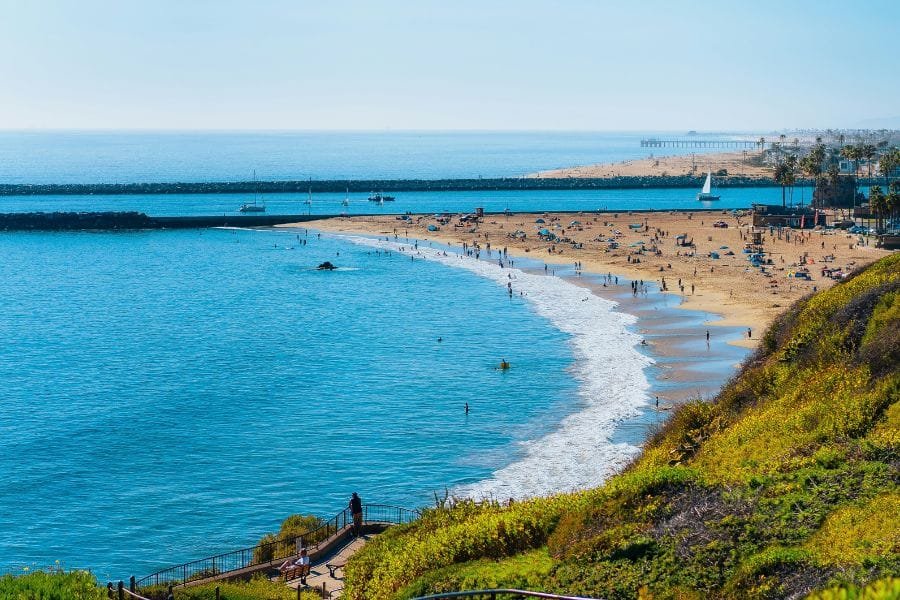 A scenic aerial view of a sunny coast showcasing popular things to do in Newport Beach, with beachgoers enjoying the clear ocean waters, sandy shore, and grassy overlook.