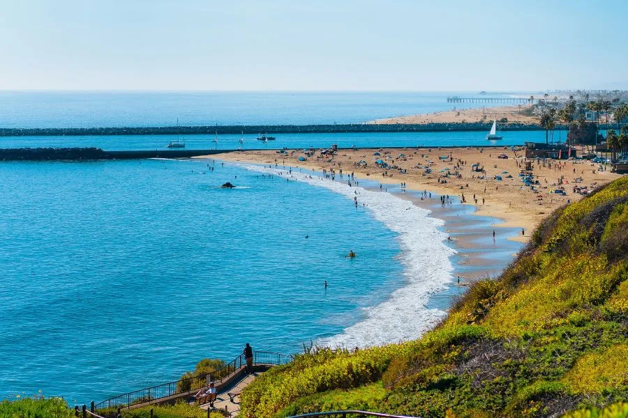 A scenic aerial view of a sunny coast showcasing popular things to do in Newport Beach, with beachgoers enjoying the clear ocean waters, sandy shore, and grassy overlook.