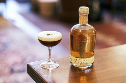 A freshly prepared Mexican Coffee Cocktail in a coupe glass with a smooth foam top and coffee beans, next to a bottle of artisanal mezcal on a wooden table.