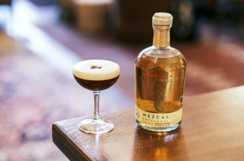 A freshly prepared Mexican Coffee Cocktail in a coupe glass with a smooth foam top and coffee beans, next to a bottle of artisanal mezcal on a wooden table.