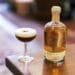 A freshly prepared Mexican Coffee Cocktail in a coupe glass with a smooth foam top and coffee beans, next to a bottle of artisanal mezcal on a wooden table.