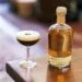 A freshly prepared Mexican Coffee Cocktail in a coupe glass with a smooth foam top and coffee beans, next to a bottle of artisanal mezcal on a wooden table.
