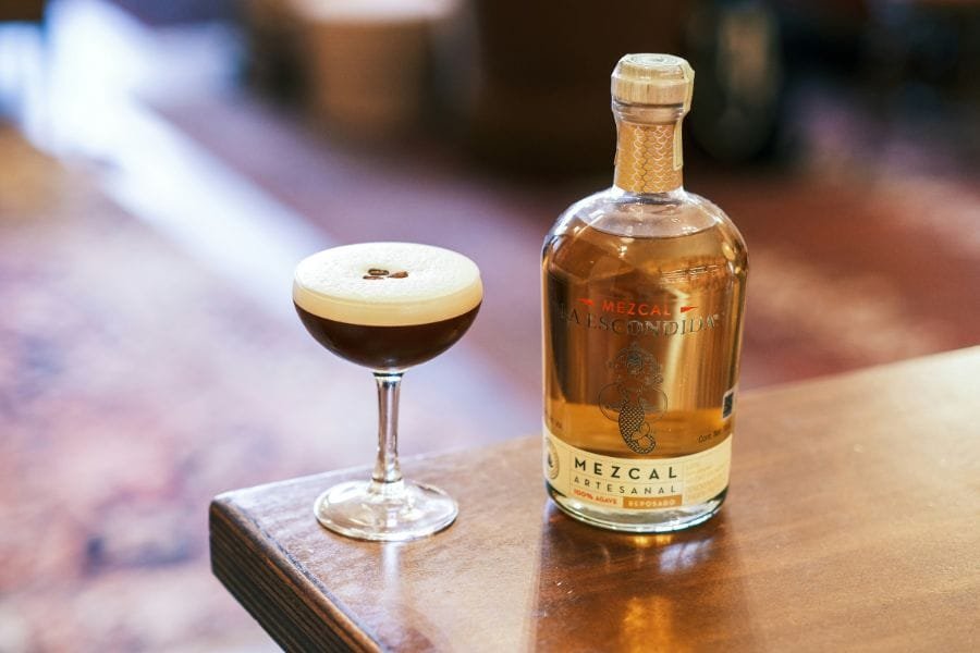 A freshly prepared Mexican Coffee Cocktail in a coupe glass with a smooth foam top and coffee beans, next to a bottle of artisanal mezcal on a wooden table.